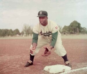 Jackie Robinson in a defensive fielding stance during practice with the Brooklyn Dodgers, wearing a vintage uniform and glove, standing at base on a dirt field in a color photograph.