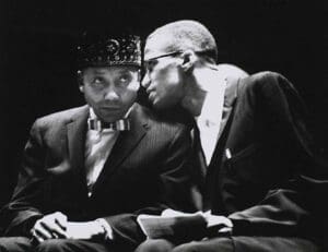 Elijah Muhammad listens as Malcolm X leans in to speak during a historic Nation of Islam event, symbolizing mentorship and deep strategic dialogue in the 1960s civil rights era.