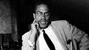 Black and white photo of Malcolm X seated in a thoughtful pose, wearing glasses and a striped suit, symbolizing his role as a bold and intellectual leader in Black American history.